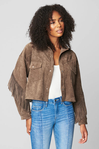 Woman wearing a brown suede jacket with fringe, white top, and blue jeans on a gray background